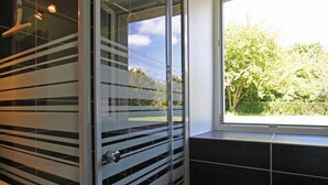 Ein Badezimmer mit Duschkabine aus Glas, einem Fenster mit Blick ins Grüne und schwarzen Fliesenwänden.