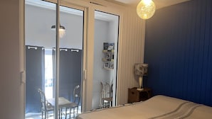 A bedroom with a bed, a nightstand, a lamp, a sliding glass door leading to a balcony, and a wall-mounted shelf with pictures.