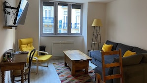 A modern living room with a sofa, a dining table, chairs, a TV mounted on the wall, and a window with a view of buildings.