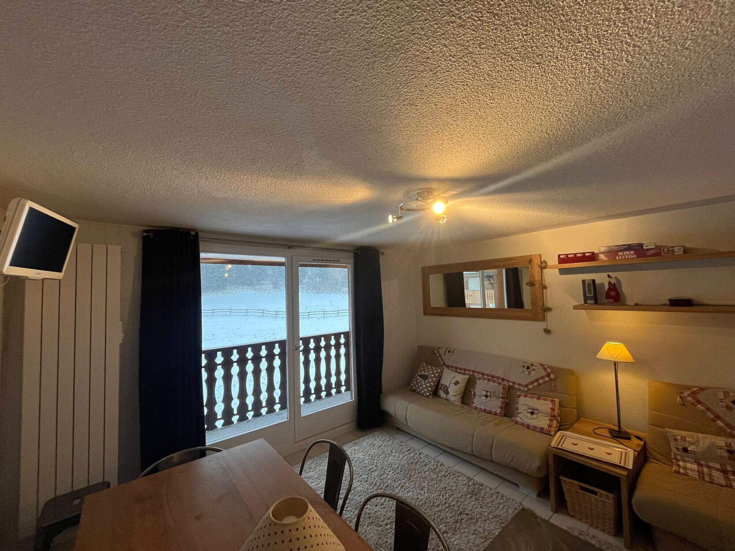 A cozy living room with a sofa, a TV mounted on the wall, a dining table, and a window with a view of a snowy landscape.