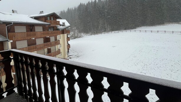 A multi-story building with balconies, a snow-covered ground, and a forested mountain in the background.