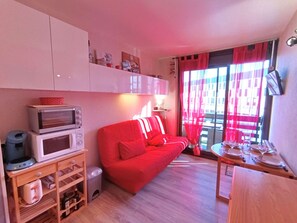 Un espacio habitable pequeño con un sofá rojo, un microondas, una mesita con vajilla y una ventana con cortinas rojas.