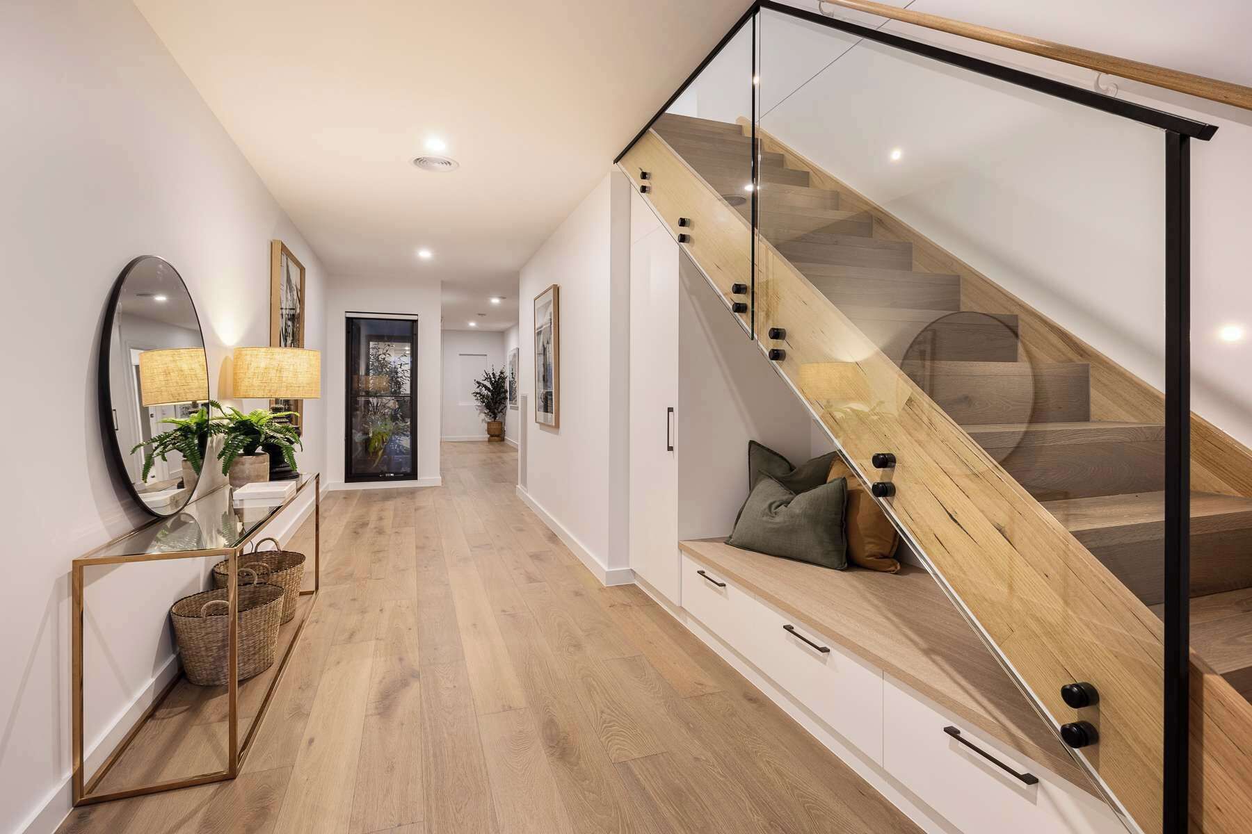 A modern interior with a wooden staircase, a glass railing, and a hallway leading to a door.