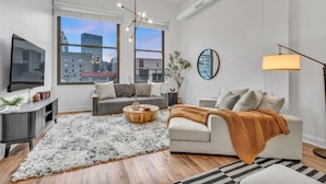 A modern living room with a sofa, coffee table, and TV, large windows with a city view, and a round mirror on the wall.