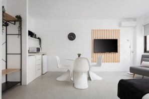 Una sala de estar moderna con comedor, muebles blancos, un televisor de pantalla plana montado en un panel de madera y un pequeño reloj en la pared.