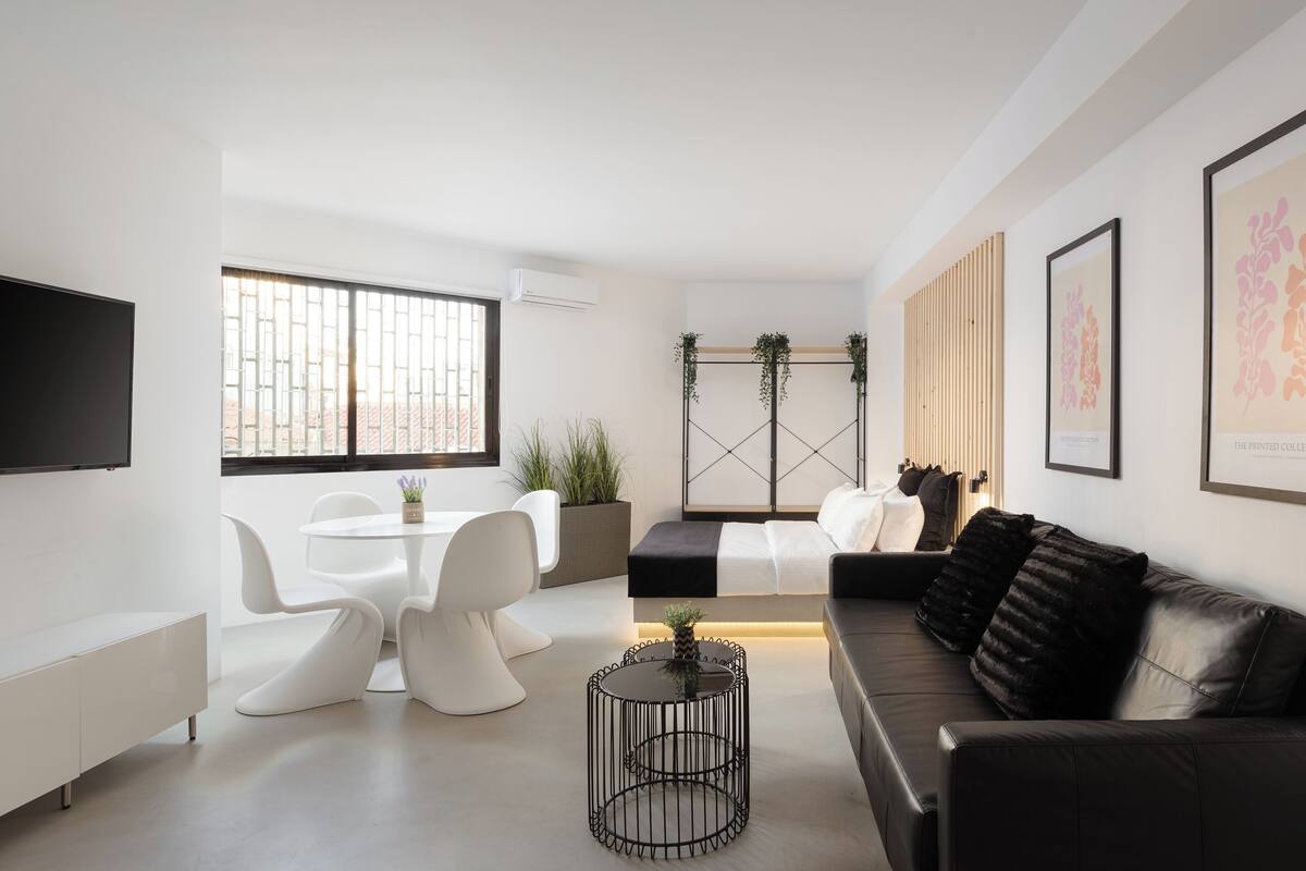 A modern living room with a black leather sofa, white chairs, a round coffee table, and a TV mounted on the wall.