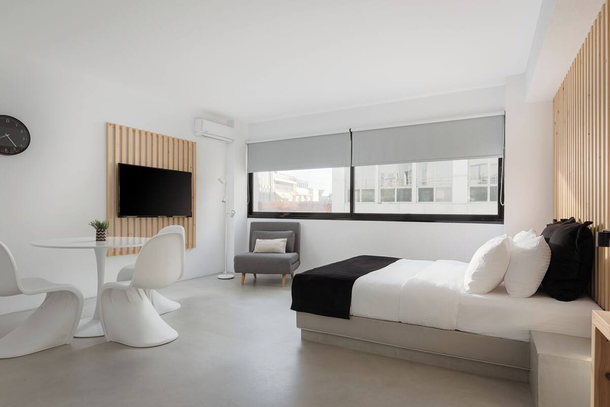 A modern hotel room with a bed, a desk, a chair, a TV, and a window with a city view.
