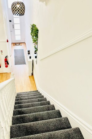 Staircase leading to an upper floor, flanked by walls and a wooden floor, with a pendant light above and a potted plant on the wall.