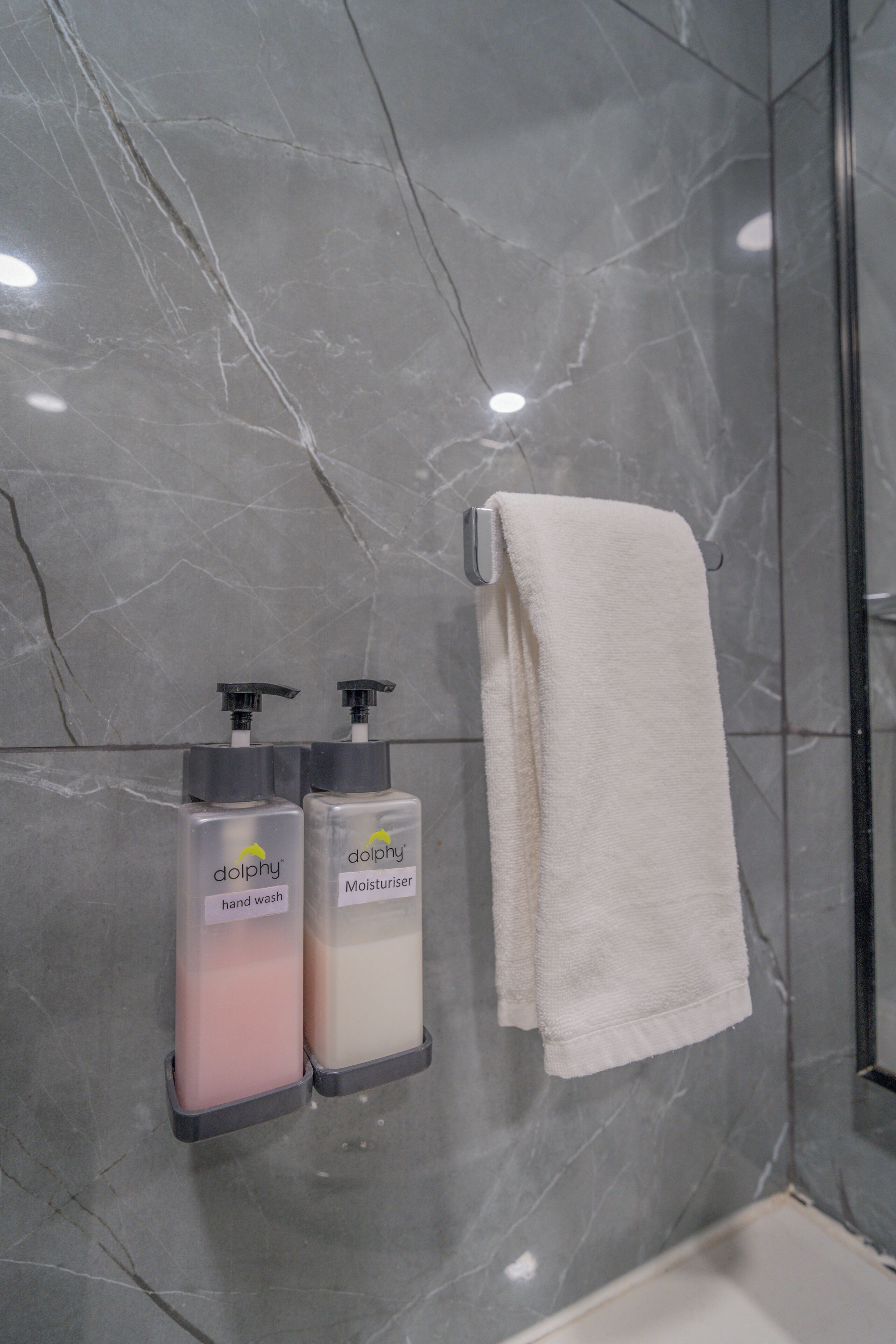 Un baño con paredes de mármol, una toalla blanca colgada en un perchero y dos botellas de jabón de manos con la etiqueta 'dolphy'.