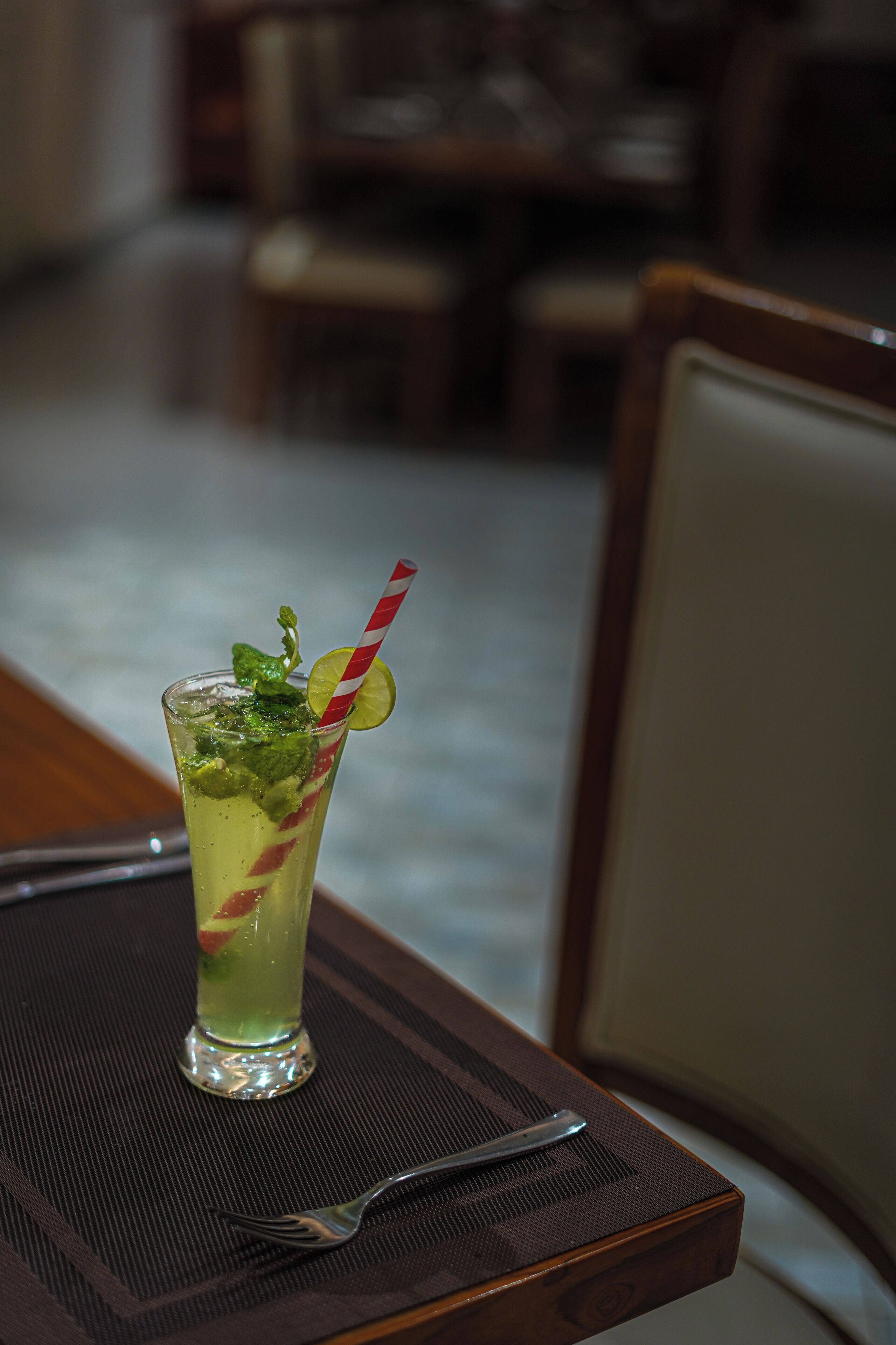 A glass of green cocktail with a lime wedge and mint garnish on a table with a fork.