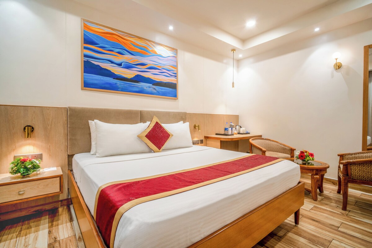 A hotel room with a large bed, a bedside table with a lamp, a small desk with a chair, and a painting on the wall.