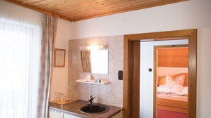 A compact bathroom with a sink, mirror, and towel rack. A wooden door leads to a bedroom with a bed.