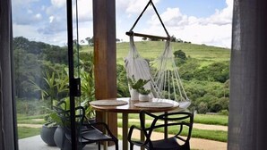 A table with chairs and a hanging chair outside, overlooking a lush green landscape. - Chalé Village Jângal (Machado)