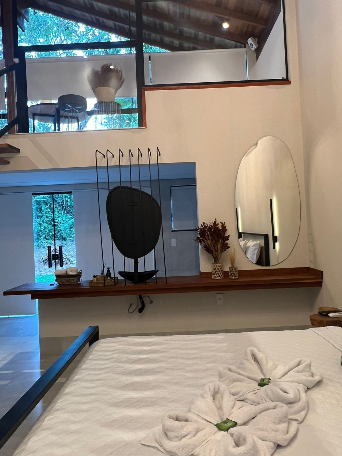 Een moderne hotelkamer met een groot bed, een houten plank met decoratieve objecten, een ronde spiegel en uitzicht op het landschap door een raam.