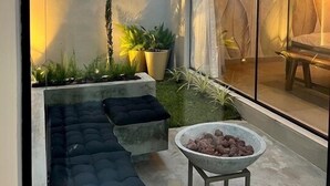 A modern outdoor seating area with a cushioned bench, a concrete planter with plants, and a stone fire pit. - Chalé Village Jângal (Machado)