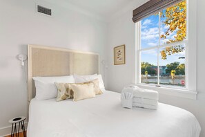 A neatly made bed with white linens and a rattan headboard, a small bedside table with a lamp, and a window with a view of trees and a stop sign.