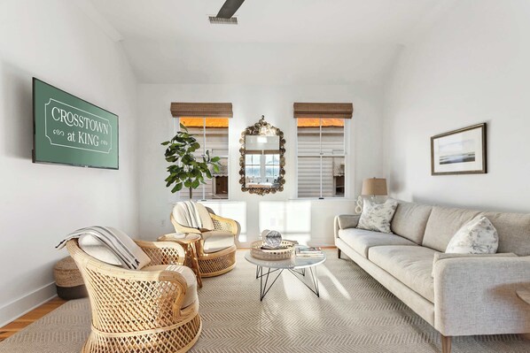 A spacious living room with a large sofa, two wicker chairs, a coffee table, and a television displaying "CROSTOWN at KING."