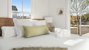 Ein Schlafzimmer mit großem Fenster, einem Bett mit weißgrünen Kissen, einer Nachttischlampe und Blick ins Freie.