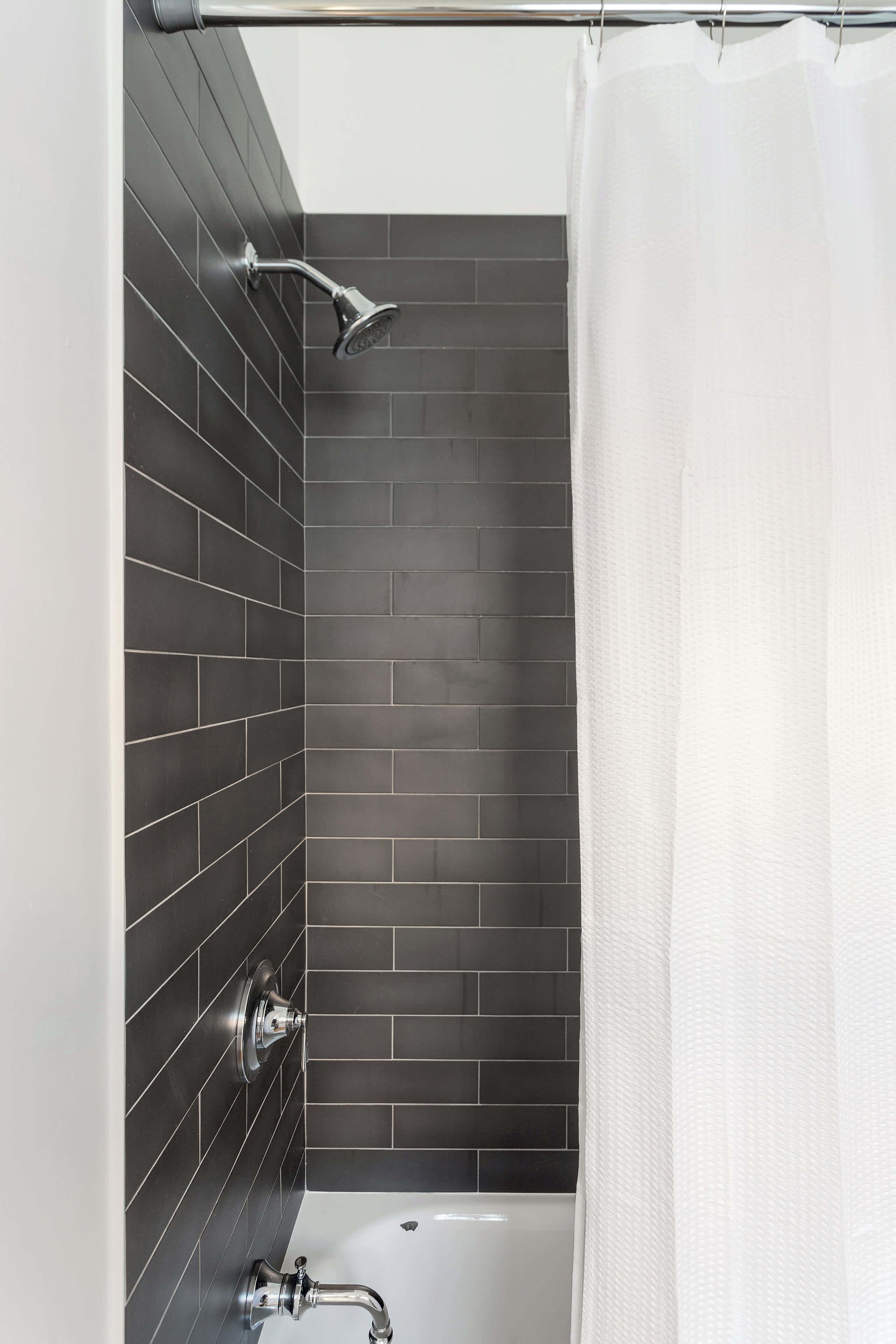 A modern bathroom with dark tiled walls, a white bathtub, and a showerhead.