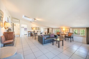 A spacious living area with a sofa, chairs, a coffee table, and a dining table. There are framed pictures on the walls and a window with curtains.