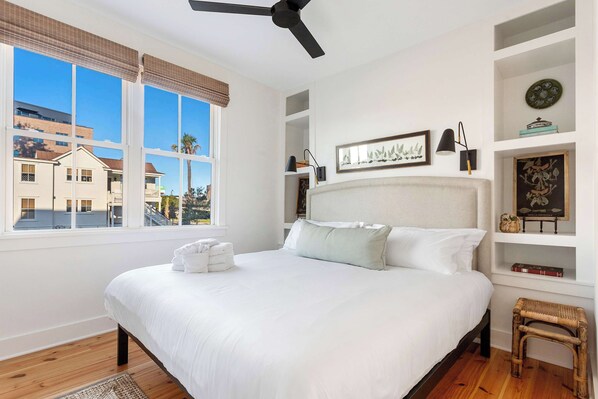 A bedroom with a large bed, a ceiling fan, and a view of buildings outside.