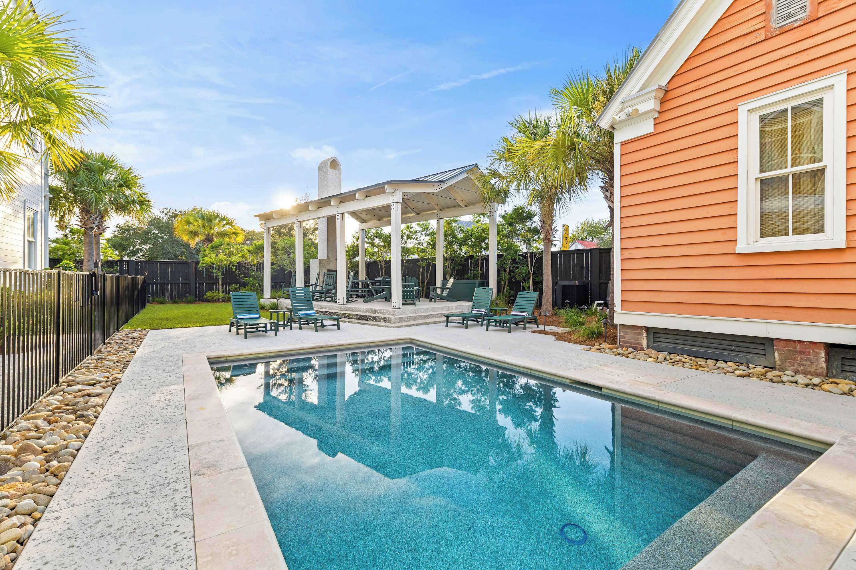 A swimming pool with a pergola and lounge chairs, surrounded by a fence and palm trees.