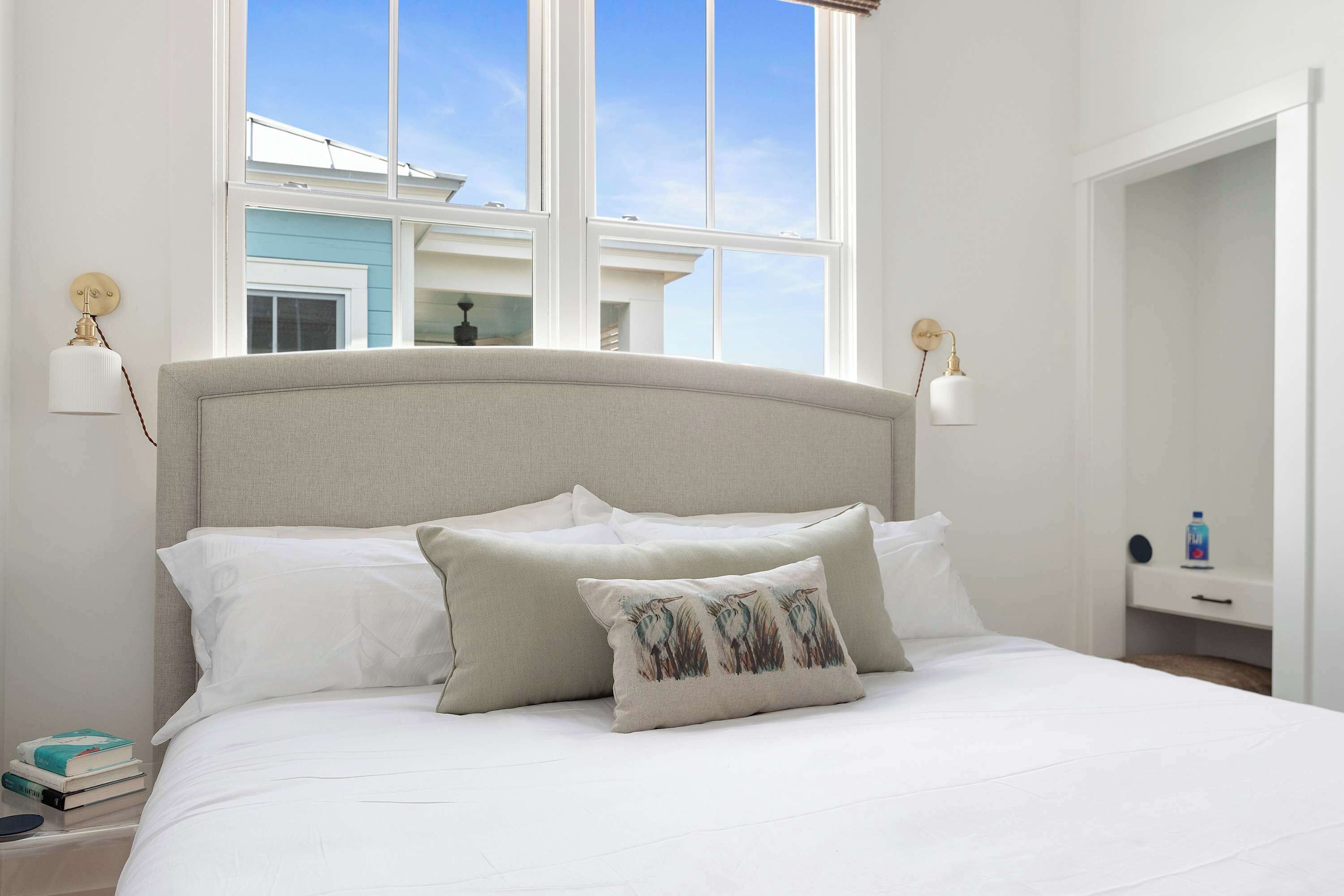 A neatly made bed with white linens and two pillows, a bedside table with a lamp, and a view of a building through large windows.