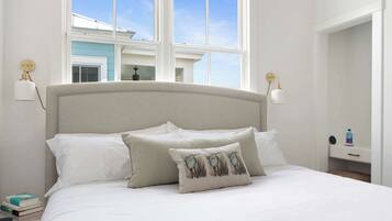 A neatly made bed with white linens and two pillows, a bedside table with a lamp, and a view of a building through large windows.