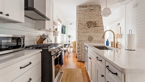 Une cuisine moderne avec des meubles blancs, un mur d’accent en briques et un îlot central doté d’un évier et d’une cuisinière.