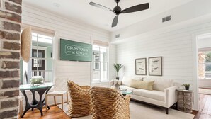 Un salon spacieux avec un canapé blanc, des poufs en osier, une table basse en verre et un ventilateur de plafond.