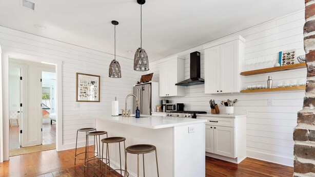 Una cocina moderna con armarios blancos, una isla central con taburetes, iluminación colgante y una pared de ladrillo decorativa.