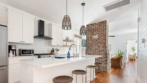 A modern kitchen with white cabinetry, a central island with stools, hanging pendant lights, and a brick accent wall. - 8BR Downtown Luxury Escape Near King (Charleston)