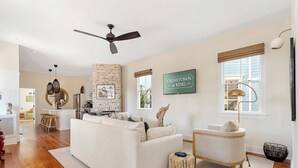 A spacious living room with a white sofa, a small round ottoman, and a vintage-style armchair. A ceiling fan and pendant lights are visible, along with a brick accent wall and a framed sign on the wall.