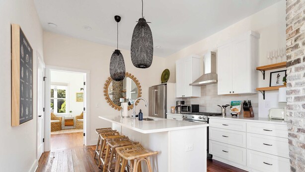 A modern kitchen with white cabinetry, a central island with stools, hanging pendant lights, and a brick accent wall.