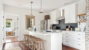 A modern kitchen with white cabinetry, a central island with bar stools, a large mirror, and a brick accent wall. - Downtown 3BR Home - Near King Street! (Charleston)
