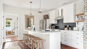 Una cocina moderna con armarios blancos, una isla central con taburetes y un espejo grande, además de un muro decorado con ladrillos.