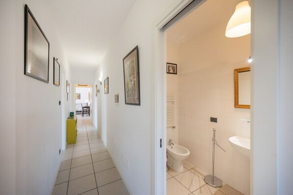 A hallway with framed pictures, a yellow cabinet, and a bathroom with a bidet, sink, and mirror.
