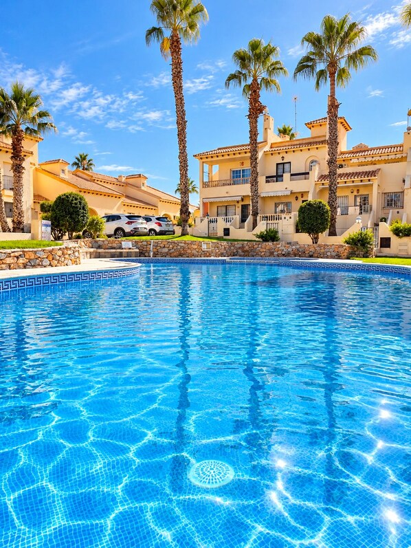 Una piscina con agua azul cristalina, rodeada de palmeras y edificios de arquitectura mediterránea.