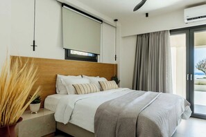 Ein modernes Schlafzimmer mit einem Holz-Kopfteil, einem großen Bett mit weißer Bettwäsche und einer grauen Decke, einer Topfpflanze und Blick ins Freie durch eine Schiebetür.