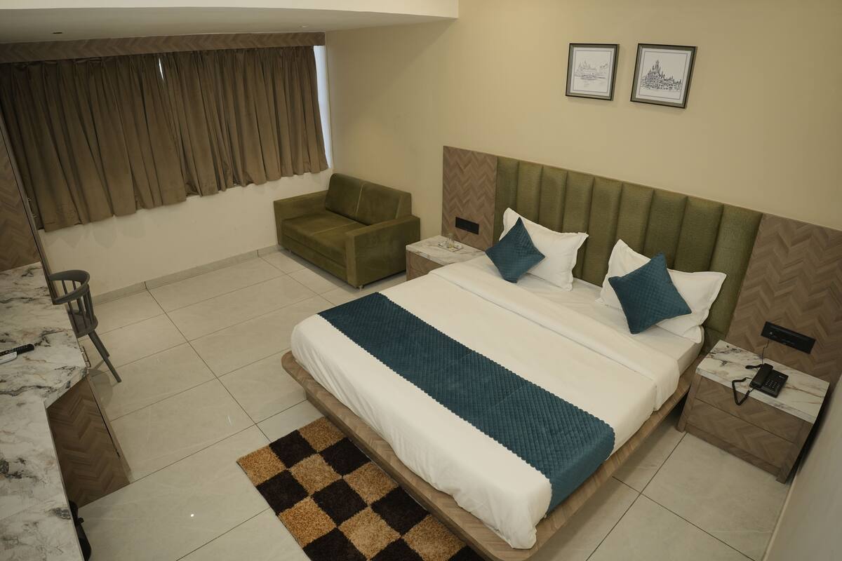 Quarto de hotel com uma cama grande, uma poltrona verde, uma mesa pequena e uma escrivaninha com cadeira. Há duas fotos emolduradas na parede.