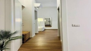 A hallway with wooden flooring, white walls, and a door at the end leading to a room with a bed and a lamp.