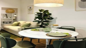 A dining area with a round table, chairs, a hanging lamp, a potted plant, and a sofa in the background.