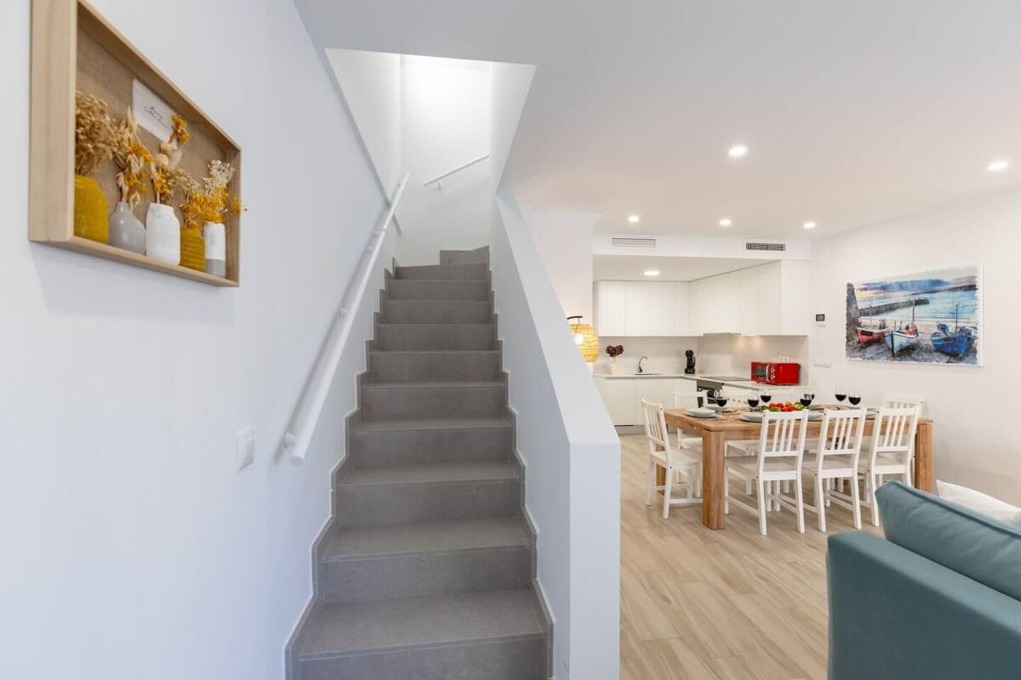 A staircase leading to a dining area with a table and chairs, a kitchen in the background, and a painting on the wall.