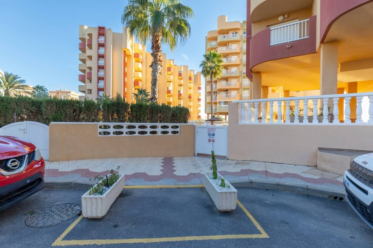 A parking area with two cars, a gated entrance, and a building with balconies and a palm tree.