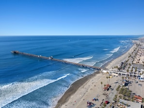 A coastal beach with a pier extending into the ocean, a sandy shore with palm trees, and a clear blue sky.