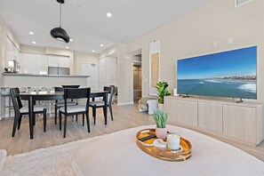 A modern living room with a large flat-screen TV displaying a coastal scene, a dining area with a wooden table and chairs, and a cozy seating area with a rug and a tray of items.