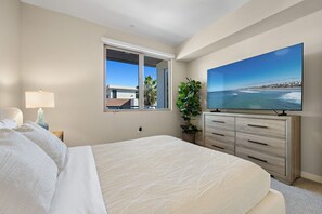 Une chambre avec un grand lit, une télévision, une fenêtre donnant sur des immeubles et des palmiers, et une plante.