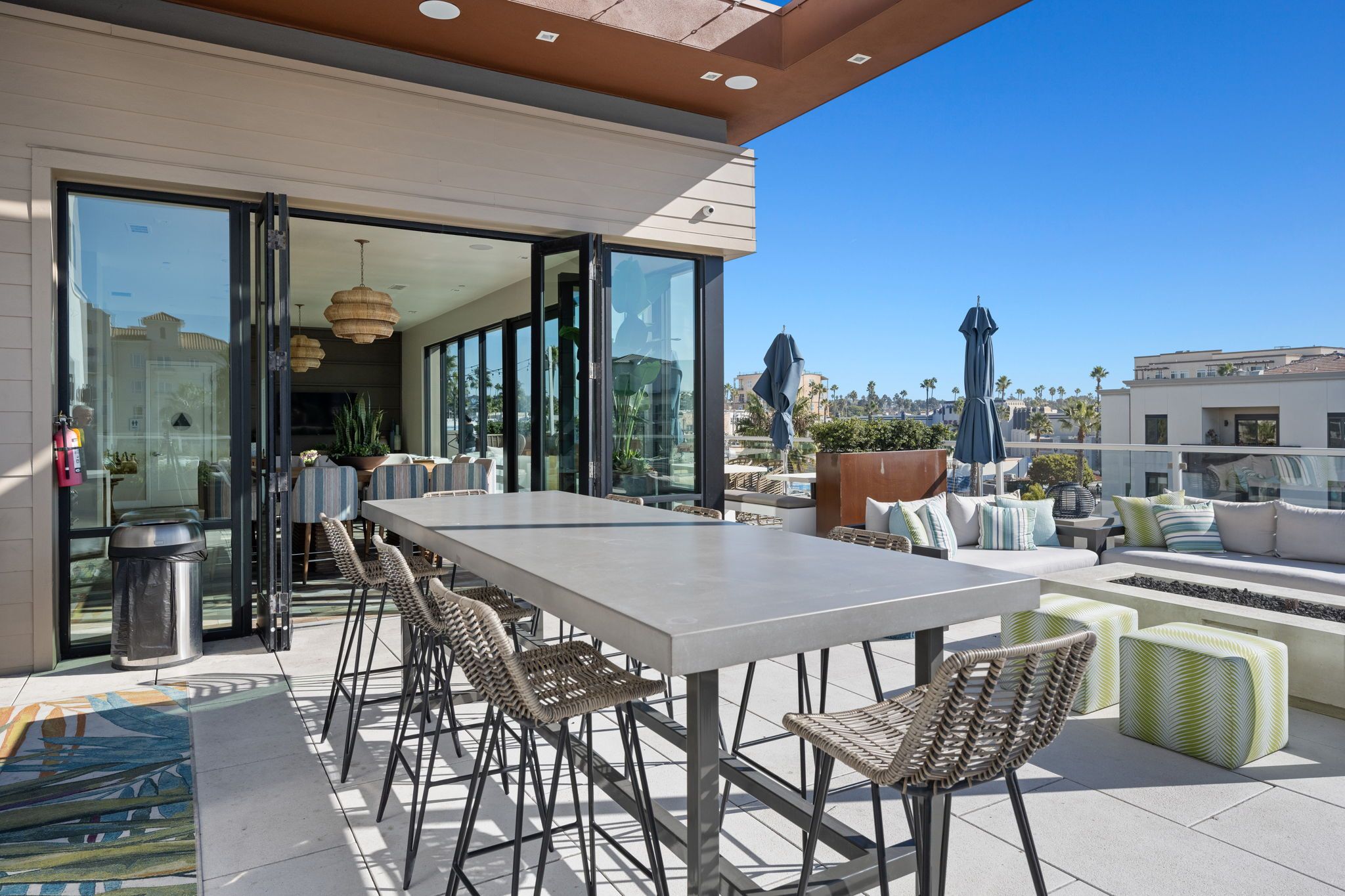 An outdoor dining area with a long table and chairs, a trash bin, and a view of the cityscape.