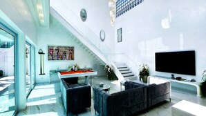 A spacious living area with a pool table, a sofa, a TV, and a staircase leading to an upper floor.