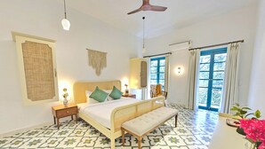 A bedroom with a bed, bedside tables, a bench, a ceiling fan, and a window with curtains. - Villa in Siolim Near Morjim Beach (Siolim)